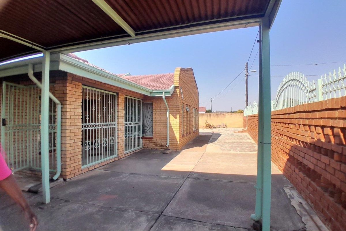 Rear exterior view of a face brick house featuring a paved courtyard, metal carport structure, and secure window bars.