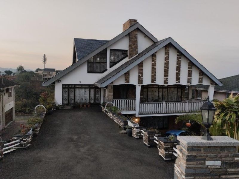 Front elevation of a multi-level house featuring a stone-clad chimney, covered balcony, and an asphalt driveway with stone-accented retaining walls.