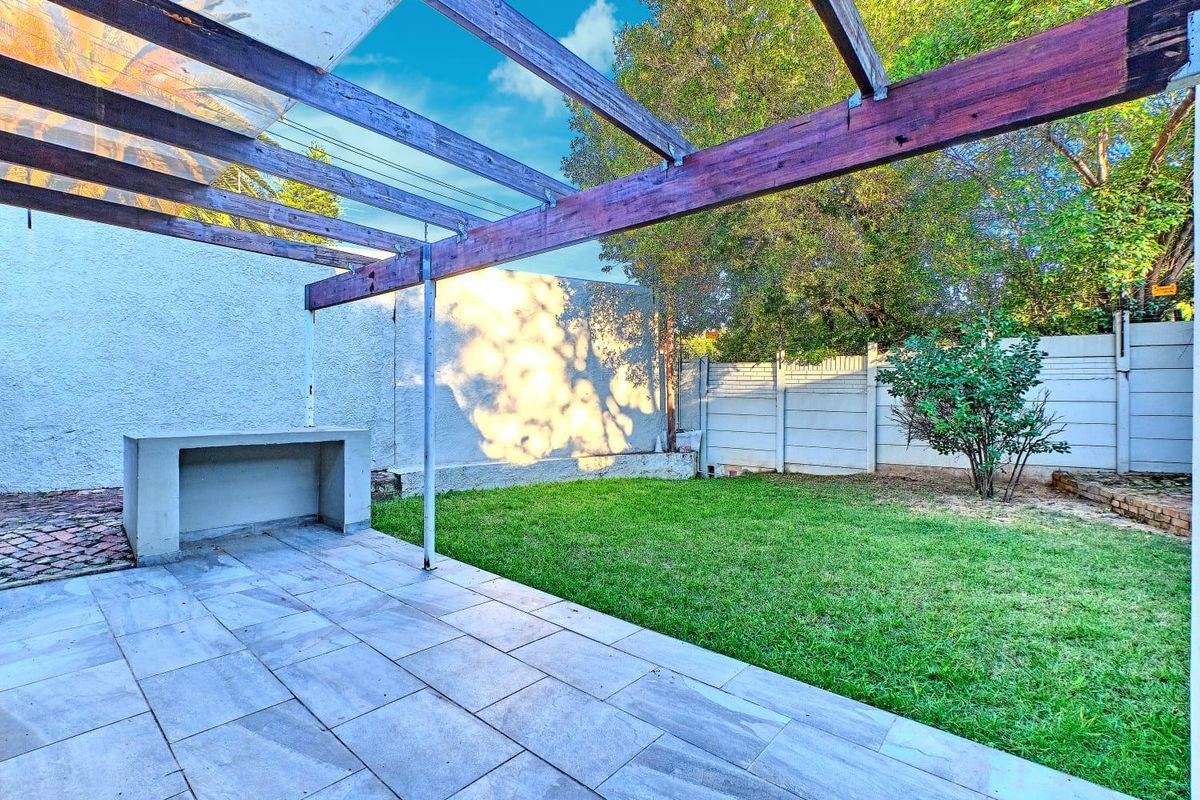 A private rear garden featuring a paved patio area with a built-in concrete counter and a timber pergola structure overlooking a small lawn.