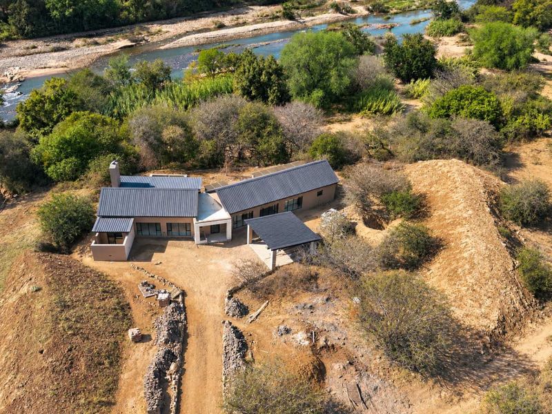 Aerial view of a modern house with a metal roof, featuring a covered patio and driveway, situated in a natural landscape near a river.