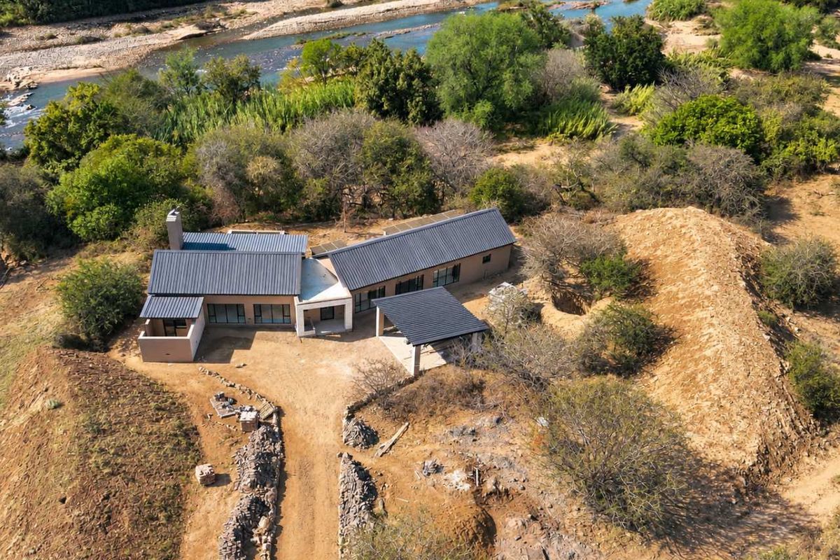 Aerial view of a modern house with a metal roof, featuring a covered patio and driveway, situated in a natural landscape near a river.