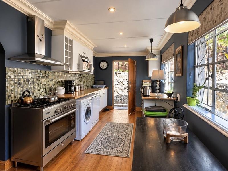 A well-appointed kitchen featuring dark blue walls, hardwood flooring, stainless steel appliances, mosaic tile backsplash, and ample natural light from large steel-framed windows.