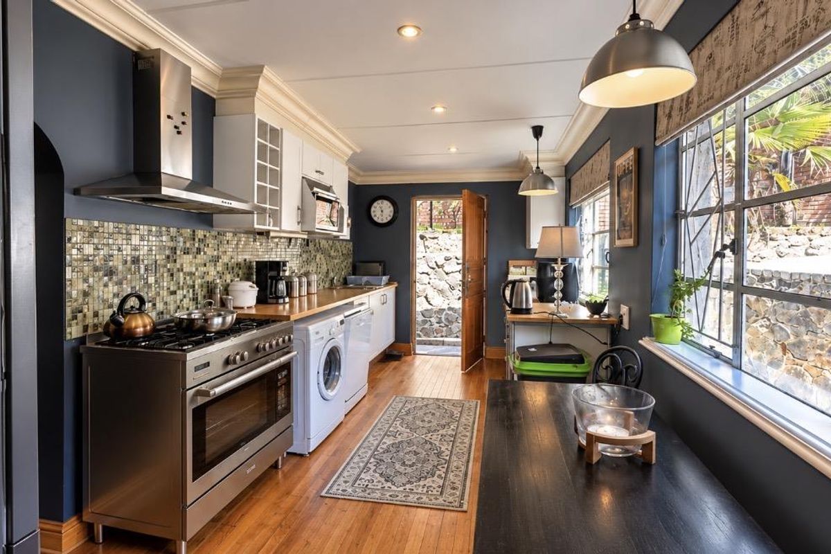 A well-appointed kitchen featuring dark blue walls, hardwood flooring, stainless steel appliances, mosaic tile backsplash, and ample natural light from large steel-framed windows.