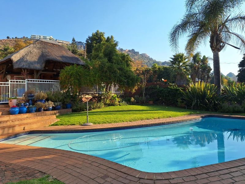 A well-maintained outdoor pool area featuring a swimming pool with brick coping, a thatched lapa, and landscaped garden surroundings.