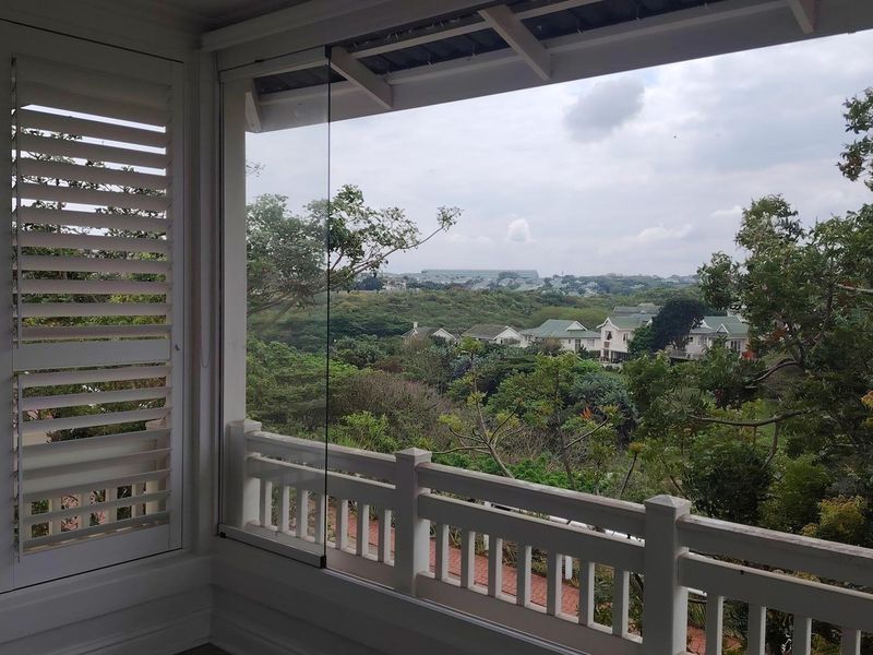 A covered balcony featuring glass sliding panels, white timber balustrades, and shutter blinds, offering an elevated view of the surrounding landscape.