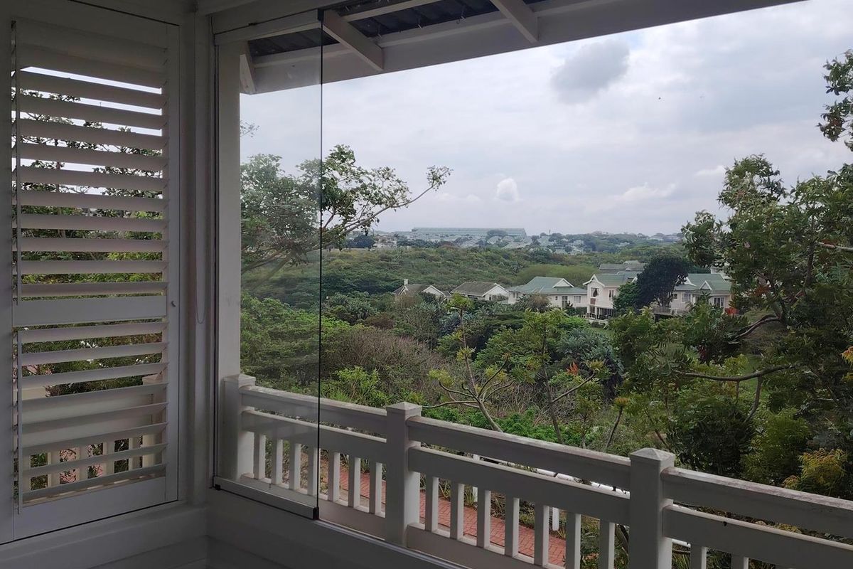 A covered balcony featuring glass sliding panels, white timber balustrades, and shutter blinds, offering an elevated view of the surrounding landscape.