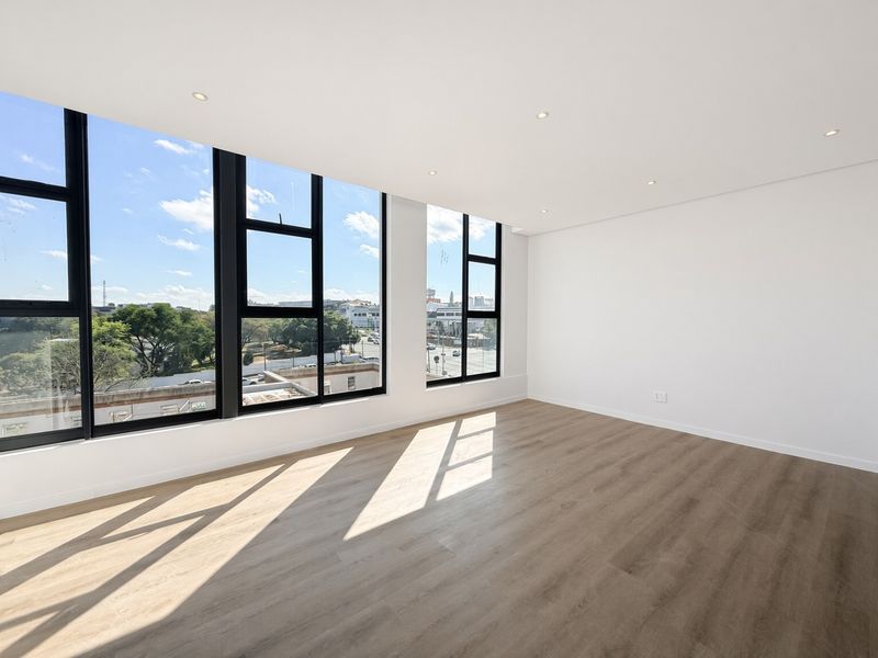 A bright, modern, and empty living area featuring large black-framed windows, recessed downlights, and high-quality laminate wood flooring with an urban view.