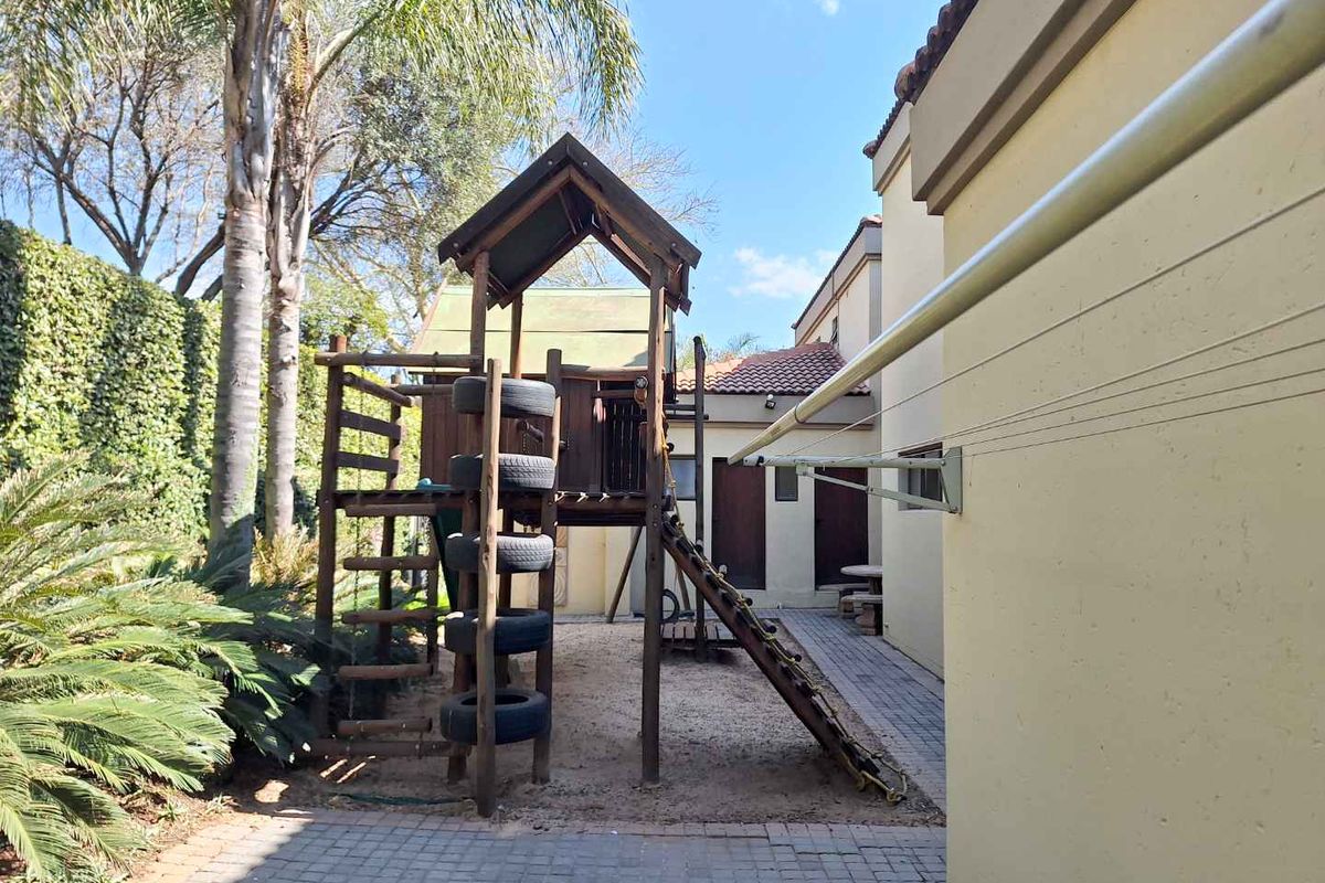 A rear garden area featuring a paved walkway, established landscaping, and a wooden play structure set against the side of the house.