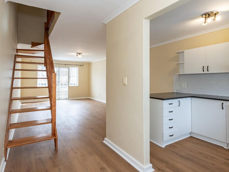 Open plan living area featuring laminate flooring, a wooden staircase, and a compact kitchen with white cabinetry and dark countertops.
