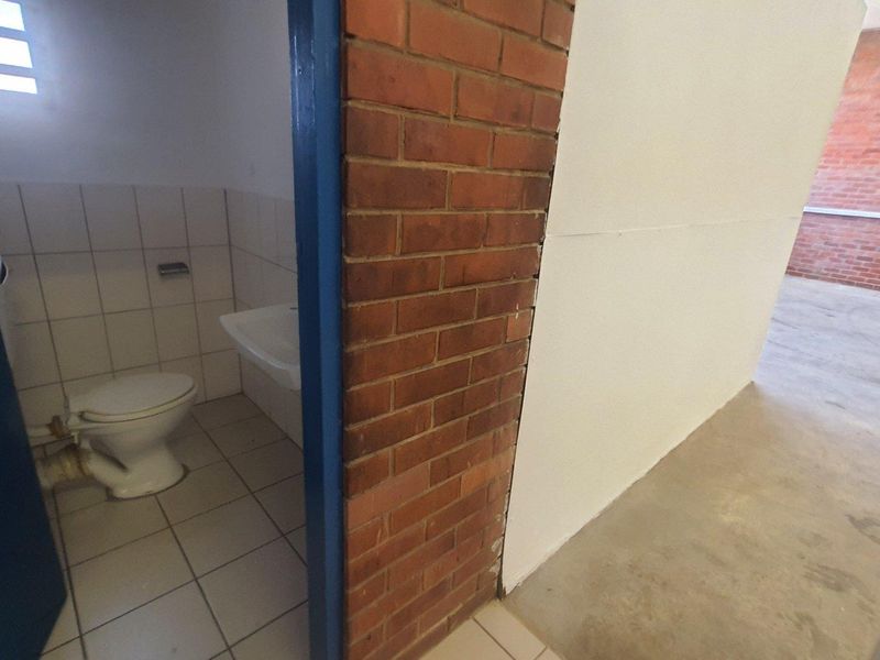 A functional industrial bathroom featuring ceramic tiling and a basic wash basin, adjacent to an open-plan area with exposed brick walls and concrete flooring.