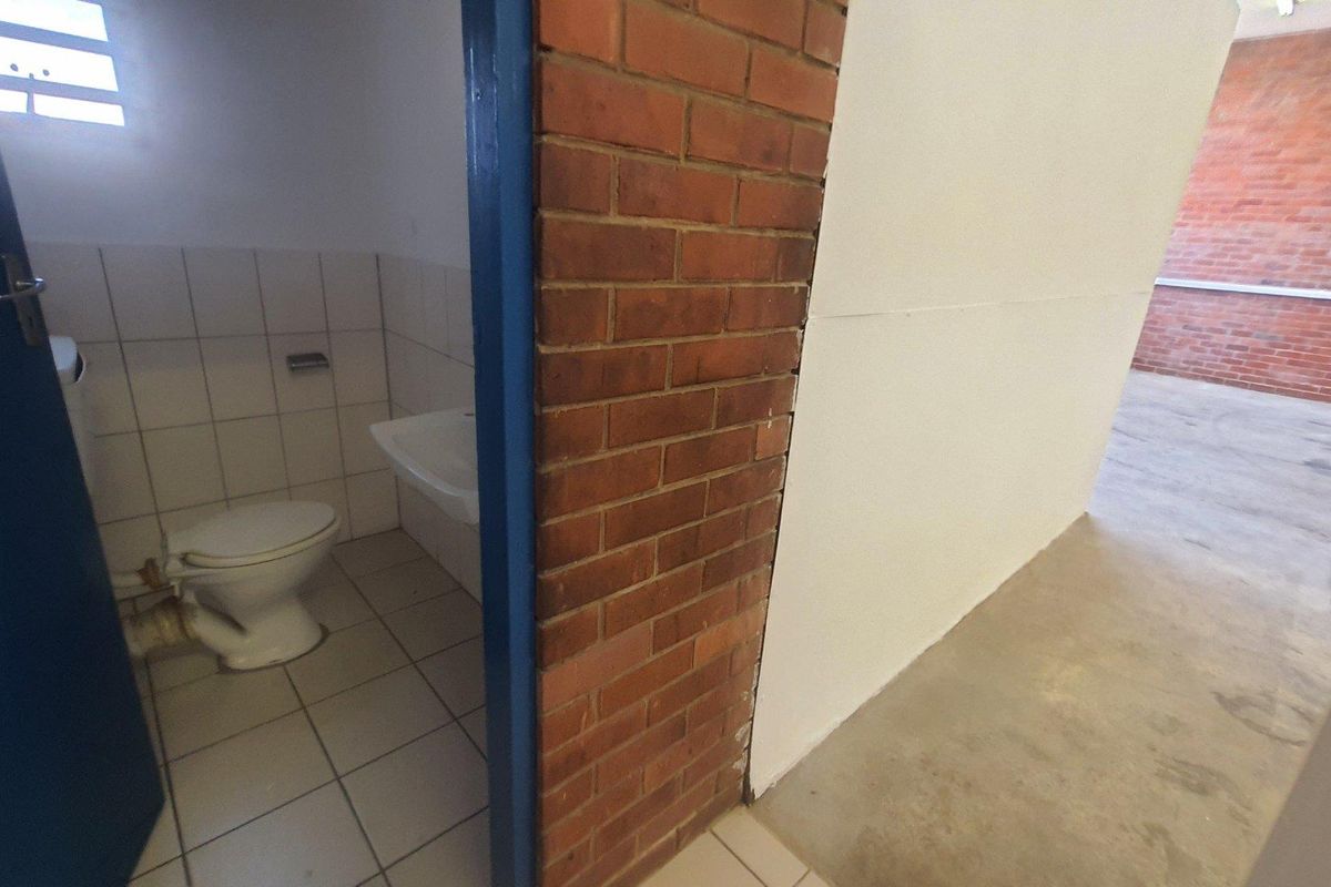A functional industrial bathroom featuring ceramic tiling and a basic wash basin, adjacent to an open-plan area with exposed brick walls and concrete flooring.