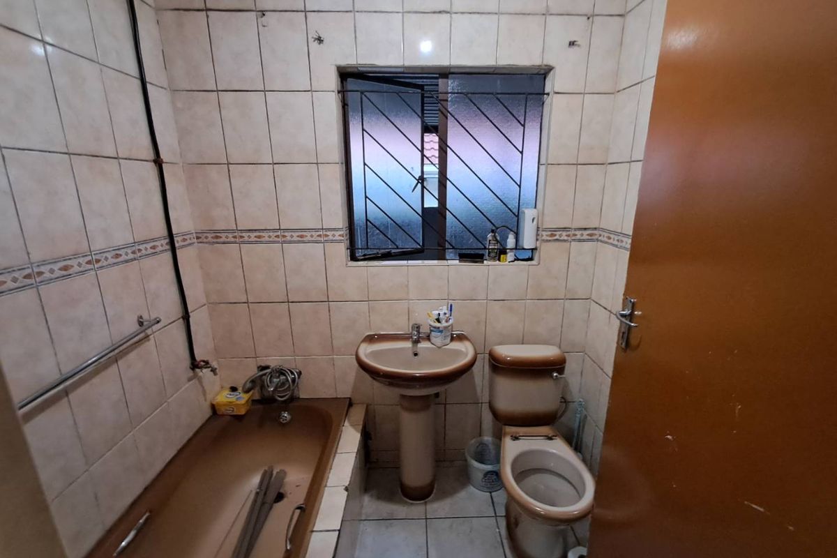 A dated bathroom featuring a brown bathtub, pedestal basin, and toilet with tiled walls and security bars on the window.