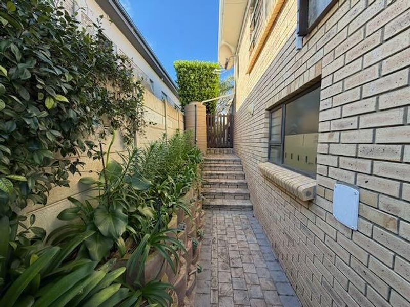 A side pathway featuring brick paving, a retaining wall with garden beds, a water tank, and a timber gate leading to the rear area.