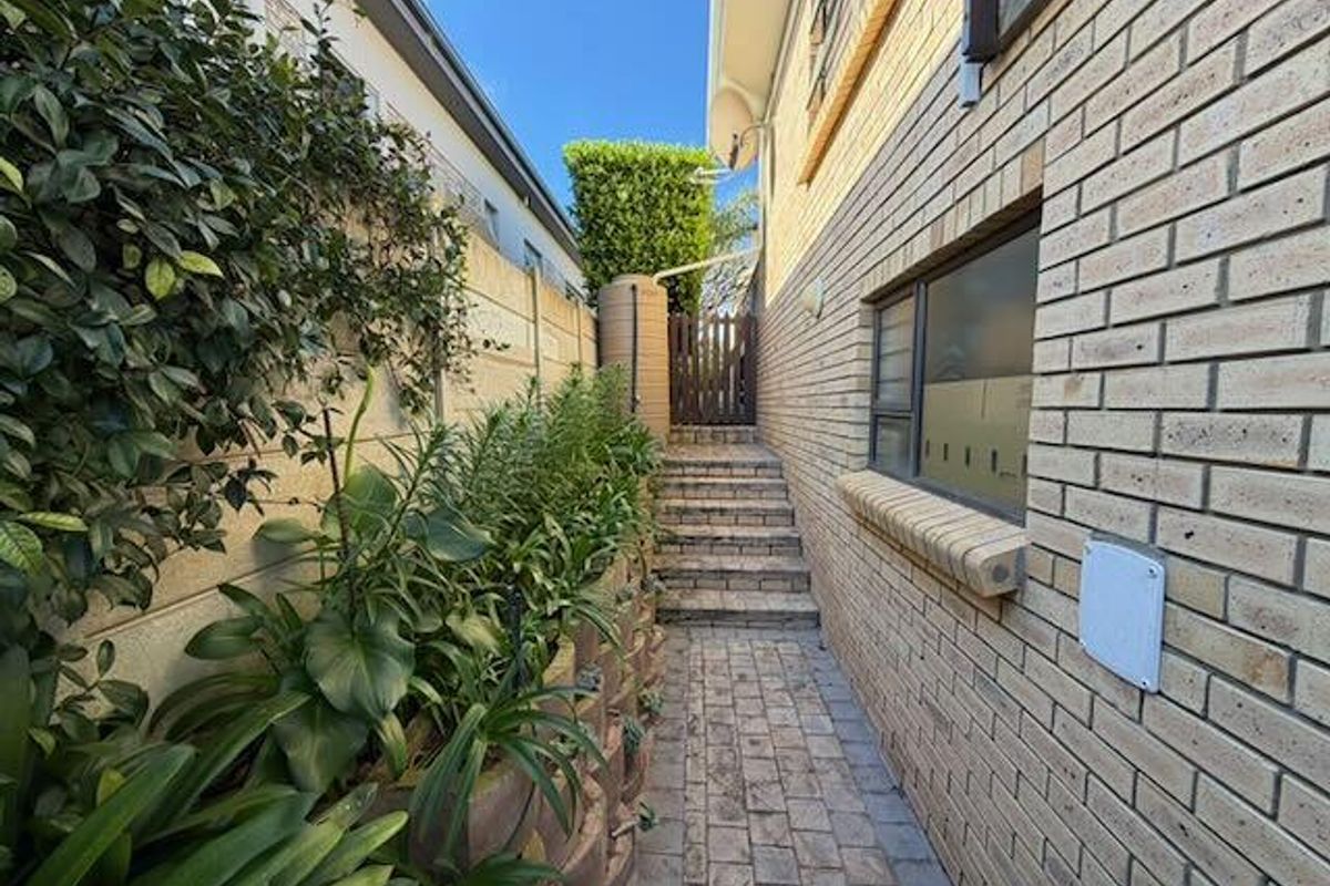 A side pathway featuring brick paving, a retaining wall with garden beds, a water tank, and a timber gate leading to the rear area.