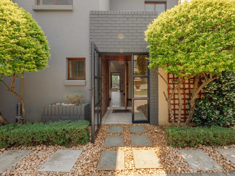 A welcoming front entrance featuring a modern steel-framed glass door, stone paver pathway, and neatly landscaped garden beds with established trees.