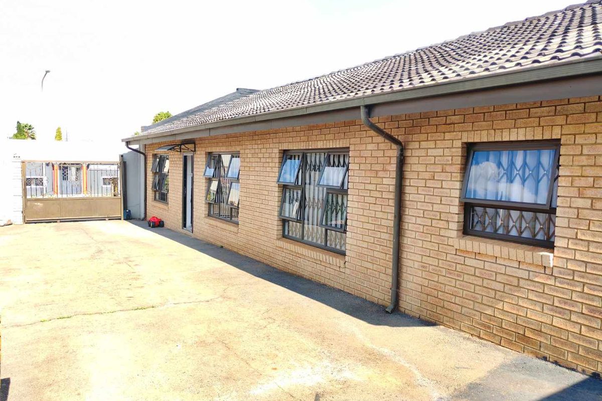 A side view of a brick house featuring security-barred windows, a tiled roof, and a concrete driveway leading to a metal gate.