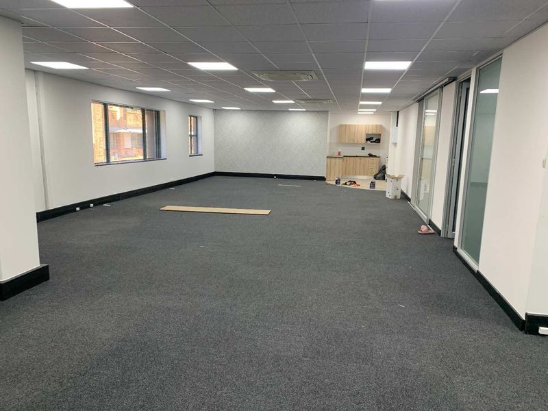 Spacious open-plan office area featuring a suspended ceiling, recessed lighting, carpeted flooring, and a small integrated kitchenette with sliding glass partitions.