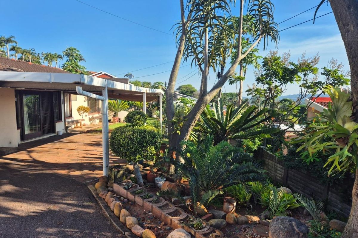 A rear exterior view featuring a covered patio area, paved driveway, and established garden landscaping with mature trees and shrubs.