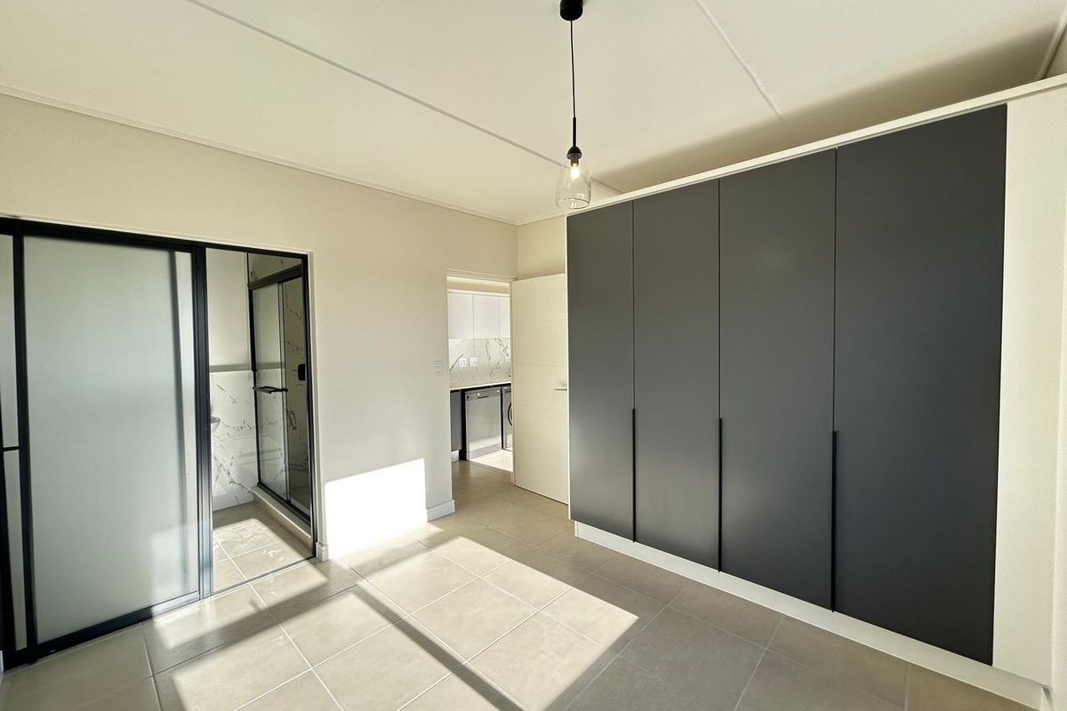 A modern bedroom featuring large built-in cupboards with dark matte doors, tiled flooring, and an en-suite bathroom with a glass-enclosed shower.