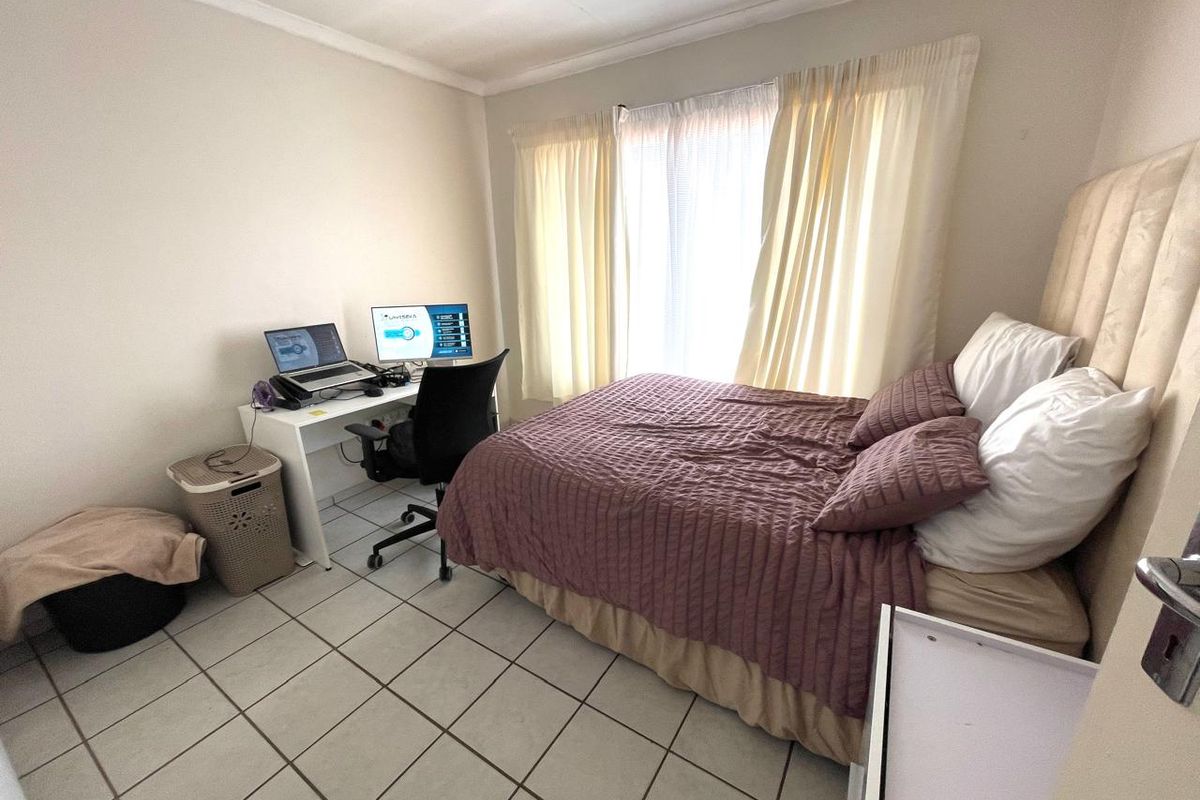 A functional bedroom featuring tiled flooring and a window with curtains, currently utilized as a combined sleeping and workspace area.