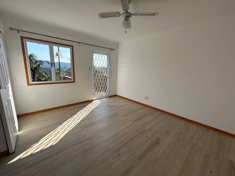 A bright, empty bedroom featuring light-toned laminate flooring, a ceiling fan, and a security gate leading to an outdoor area with scenic views.