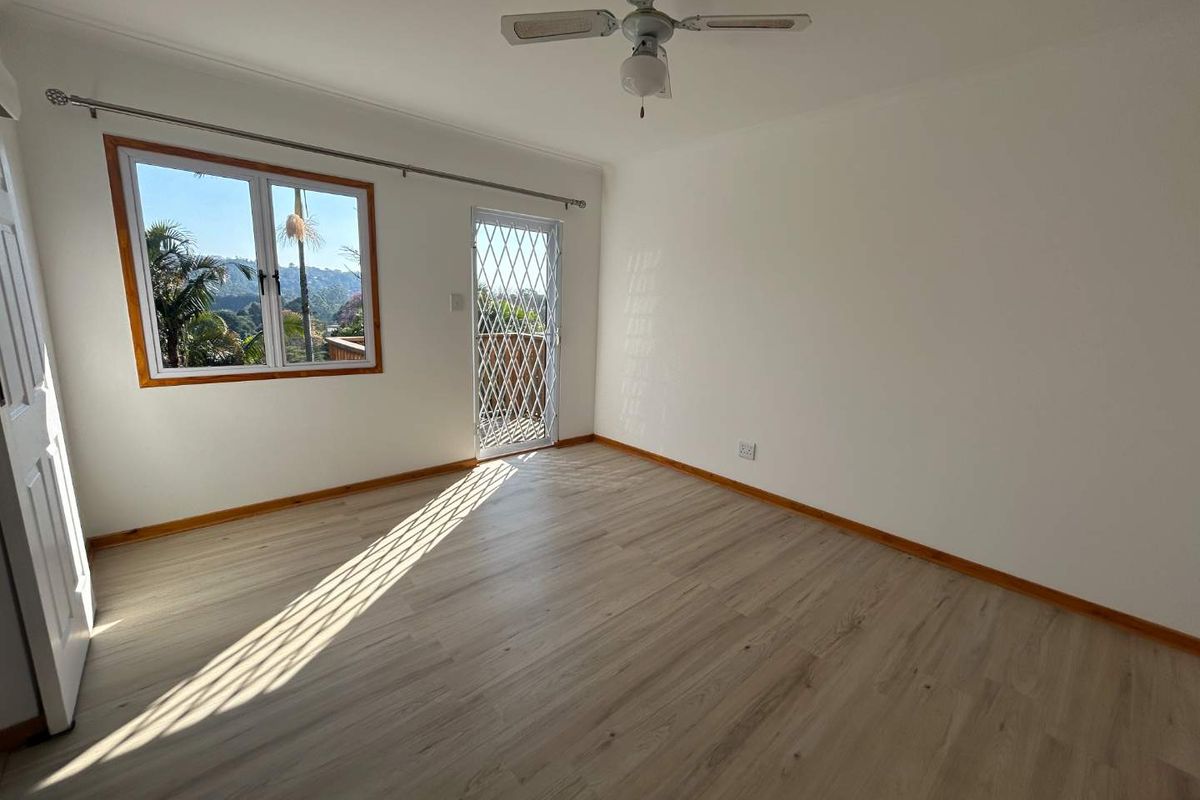 A bright, empty bedroom featuring light-toned laminate flooring, a ceiling fan, and a security gate leading to an outdoor area with scenic views.