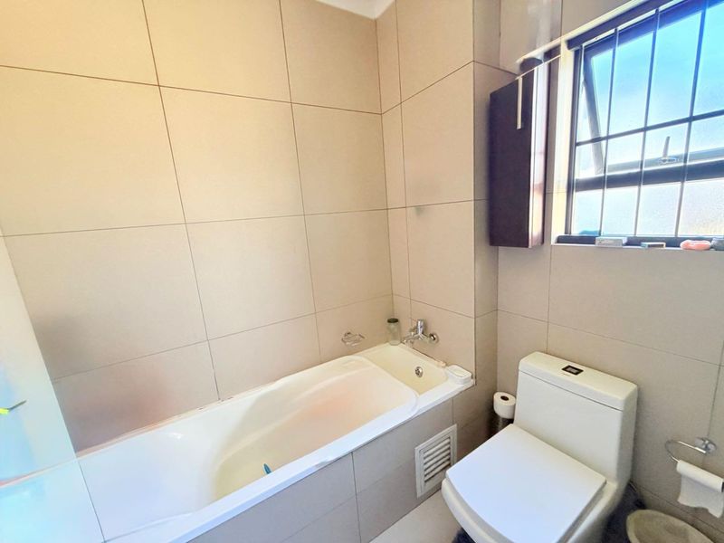 A clean, neutral-toned bathroom featuring a built-in bathtub, modern toilet, and large format wall tiles with natural light from a window.