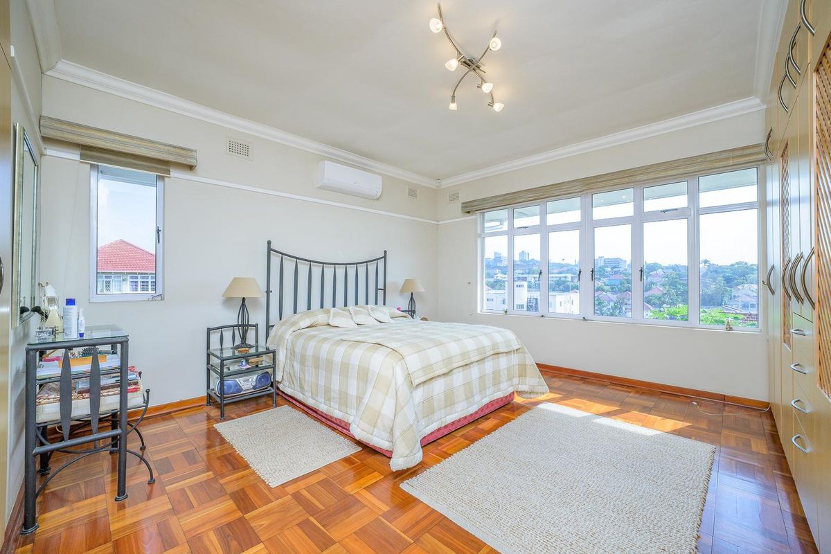 Spacious bedroom featuring polished parquet flooring, large windows with city views, built-in wooden cabinetry, and a wall-mounted air conditioning unit.