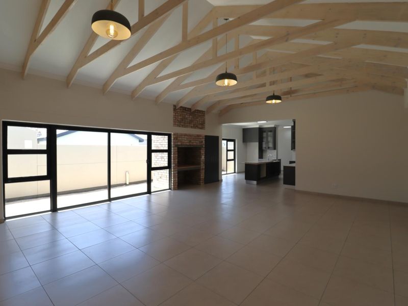 Spacious open-plan living area featuring exposed timber trusses, a brick fireplace, tiled floors, and seamless flow to the kitchen and outdoor space.