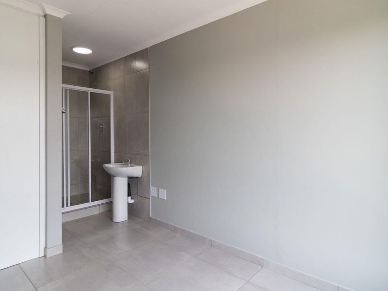 A clean, modern bathroom featuring a pedestal basin, a glass-enclosed shower, and neutral floor tiling in a well-maintained, minimalist space.