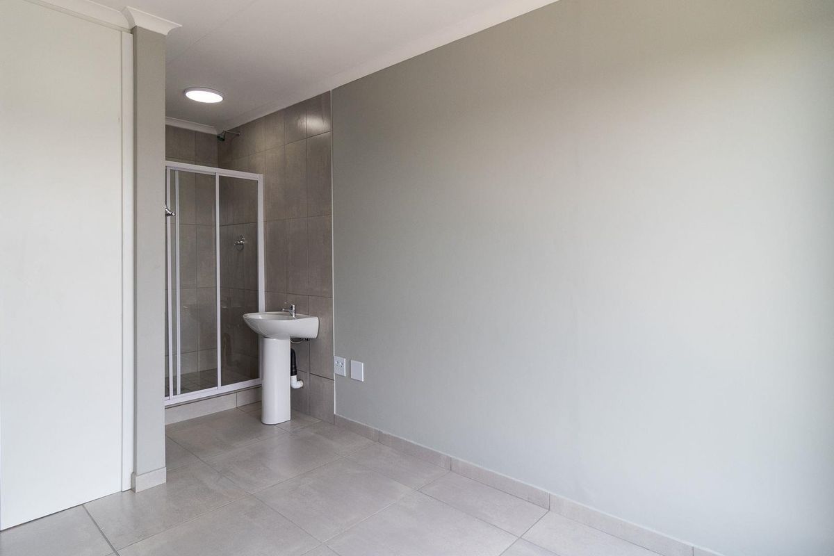 A clean, modern bathroom featuring a pedestal basin, a glass-enclosed shower, and neutral floor tiling in a well-maintained, minimalist space.