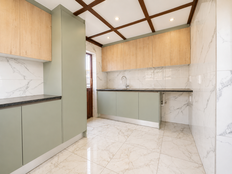 A modern, empty kitchen featuring sage green and light wood cabinetry, marble-look wall and floor tiles, and a distinctive coffered ceiling with recessed lighting.