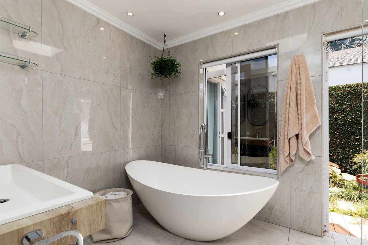 Modern bathroom featuring floor-to-ceiling marble-look tiling, a contemporary freestanding bathtub, recessed downlighting, and a sliding window providing natural light and garden views.