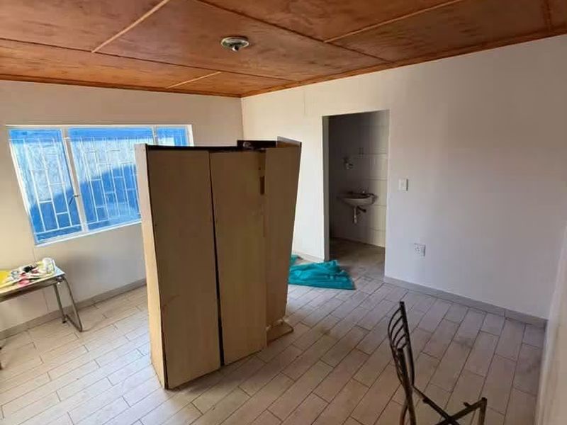 A basic bedroom featuring tiled flooring, a wooden panel ceiling, and security bars on the window, with an adjoining en-suite bathroom area.