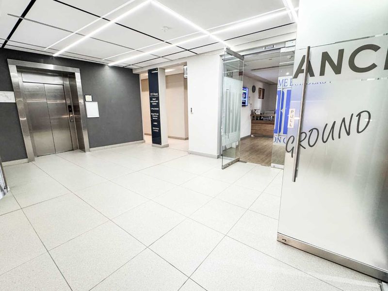 Modern commercial building lobby featuring tiled flooring, a stainless steel elevator, and glass entrance doors with clear directional signage.