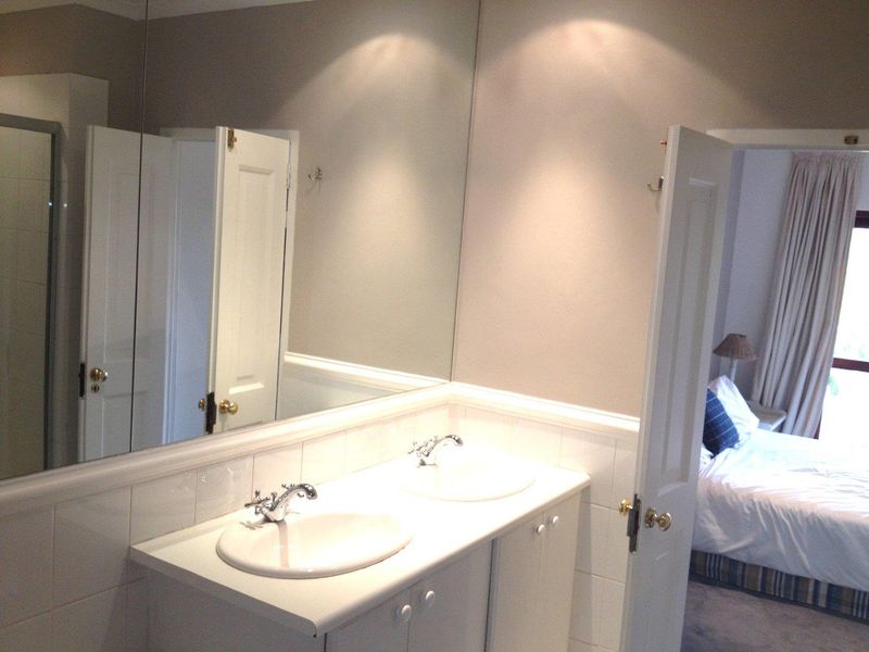 A functional bathroom featuring a double vanity with integrated sinks, large wall-to-wall mirror, and tiled splashback, adjacent to a bedroom.