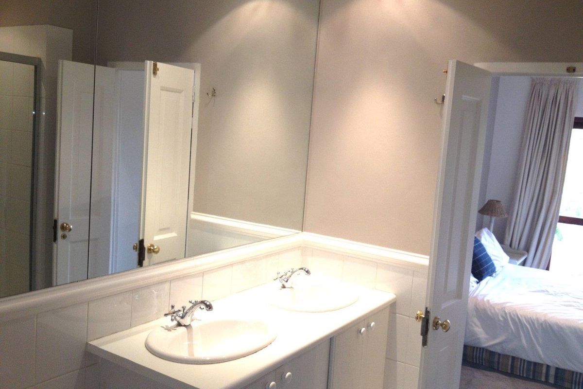 A functional bathroom featuring a double vanity with integrated sinks, large wall-to-wall mirror, and tiled splashback, adjacent to a bedroom.