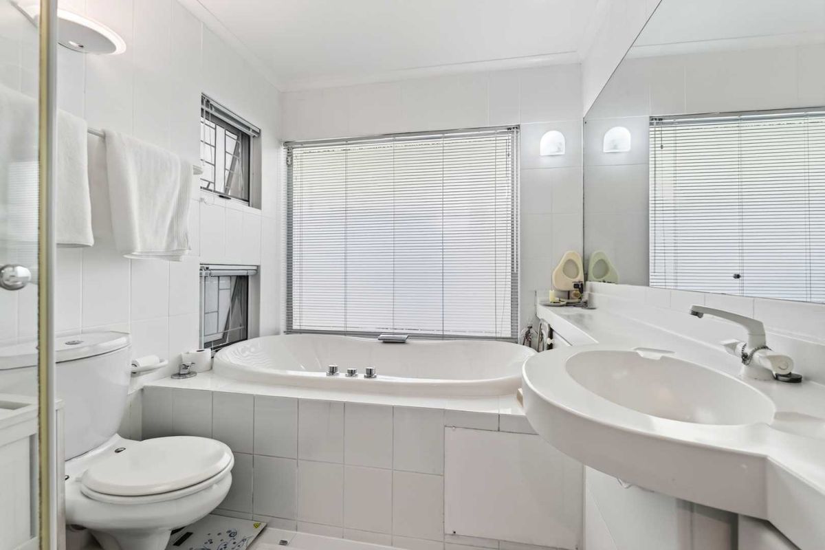 A clean, well-lit bathroom featuring a built-in bathtub, wall-mounted basin, toilet, and large wall mirror with neutral white tiling throughout.