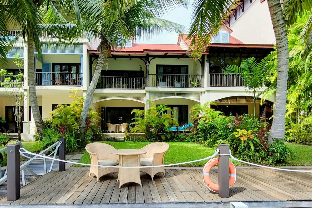 A view of the rear facade of the apartment complex, featuring balconies, ground-floor patios, and lush tropical landscaping overlooking a wooden deck area.