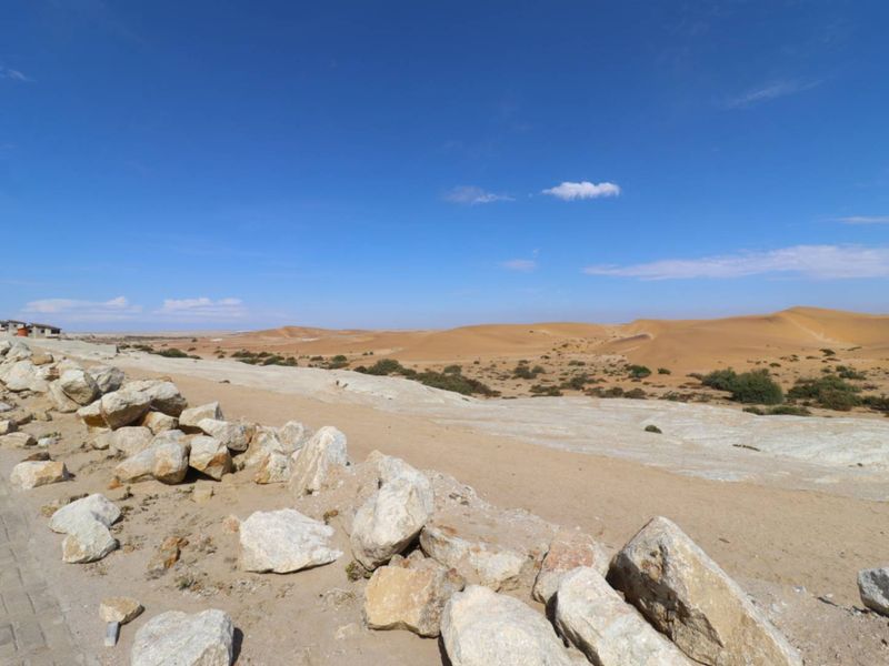A wide-angle view of the surrounding desert landscape, featuring natural sand dunes, rocky terrain, and a paved verge in the foreground.