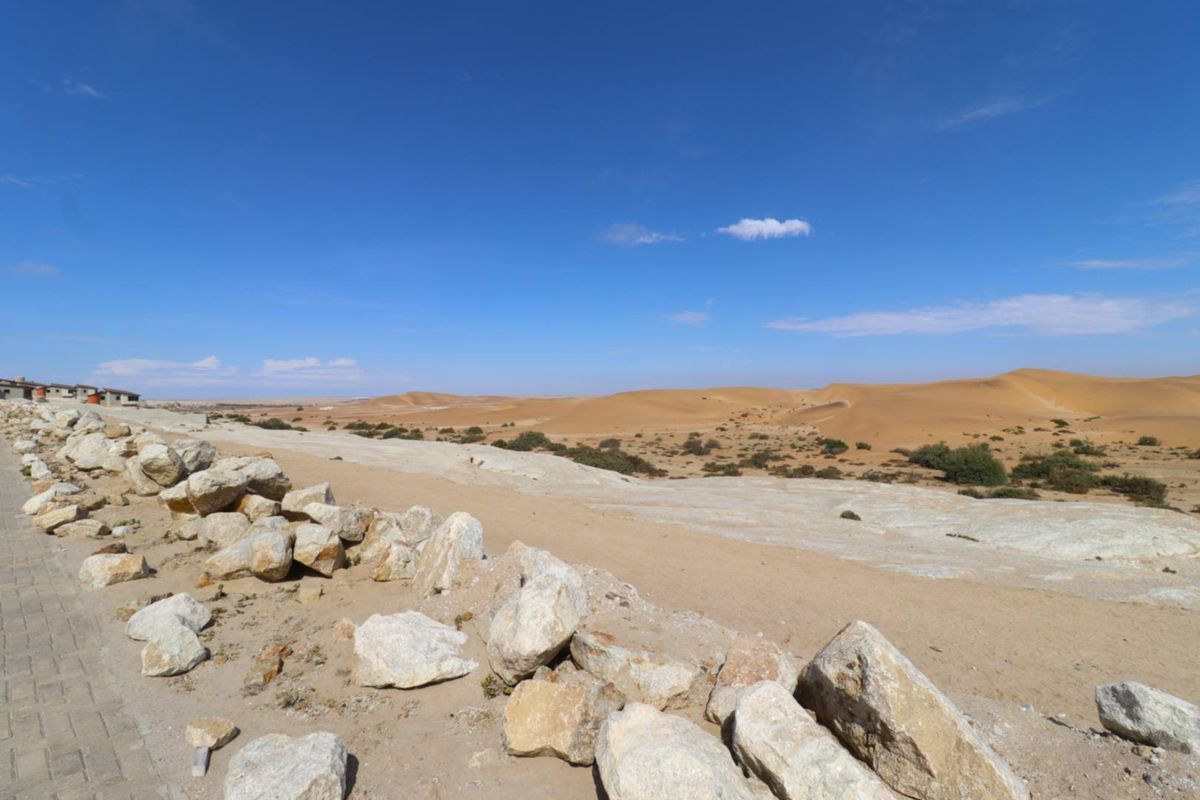 A wide-angle view of the surrounding desert landscape, featuring natural sand dunes, rocky terrain, and a paved verge in the foreground.