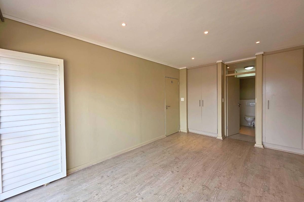 A bright, empty bedroom featuring light wood-look flooring, built-in cupboards, and an en-suite bathroom entrance.