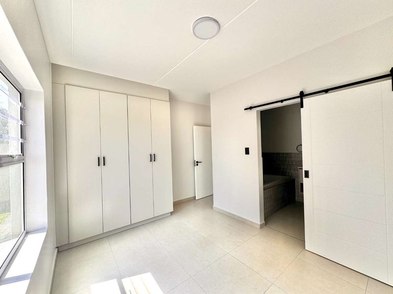 A bright, modern bedroom featuring neutral tiled flooring, ample built-in cupboard space, and a stylish sliding barn door leading to an en-suite bathroom.
