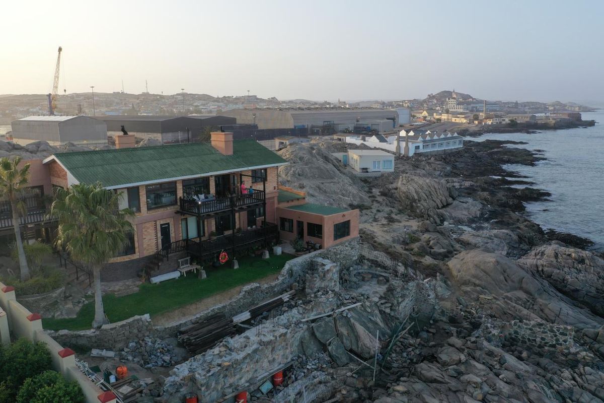 Aerial view of a coastal property featuring a multi-level house with balconies, set against a rocky shoreline and industrial harbour backdrop.