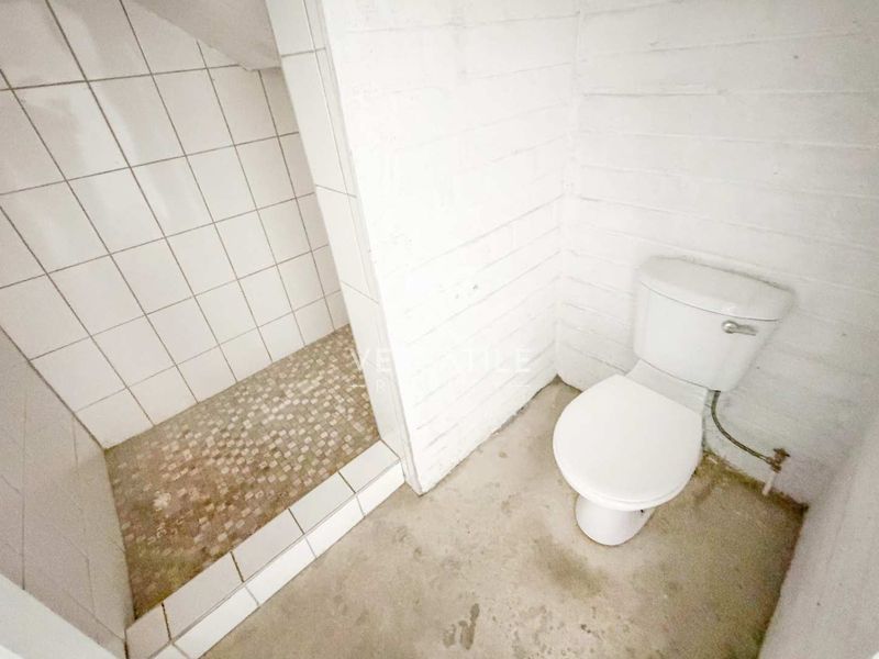 A basic industrial-style bathroom featuring a shower enclosure with tiled walls and a separate toilet, requiring cosmetic updates and floor finishing.