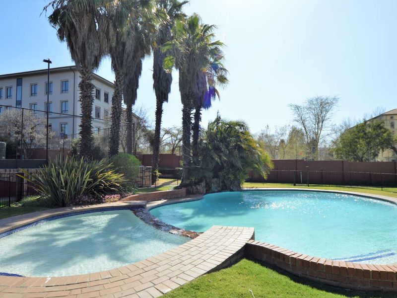 A well-maintained communal swimming pool area featuring brick coping, landscaped gardens, and mature palm trees within a secure apartment complex.