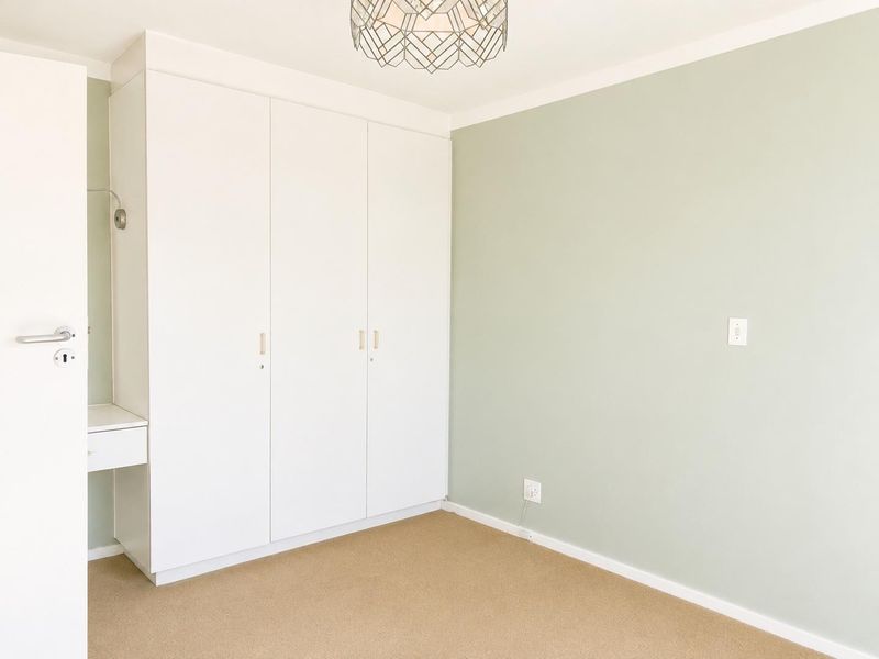 A bright, empty bedroom featuring neutral carpeted flooring, soft green walls, and a large white built-in wardrobe with an integrated vanity section.