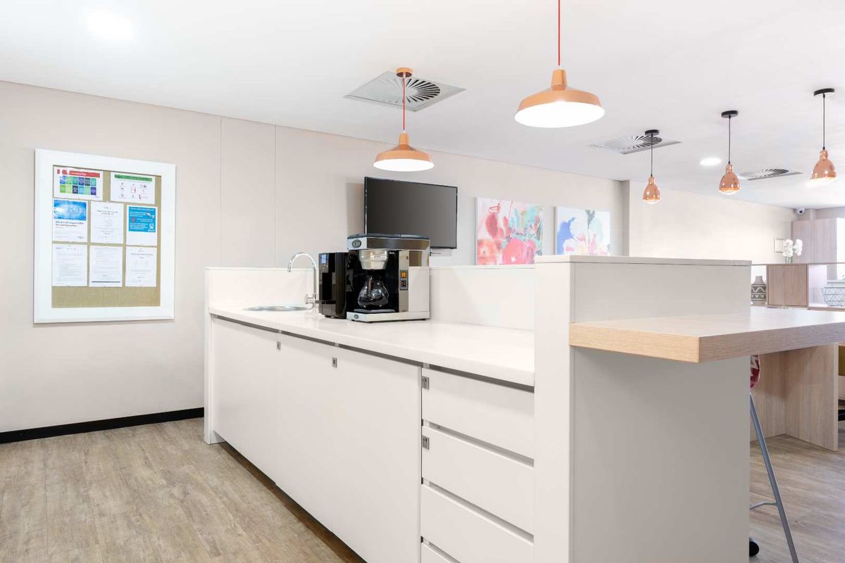 A modern office kitchenette area featuring clean white cabinetry, a built-in sink, and contemporary pendant lighting.