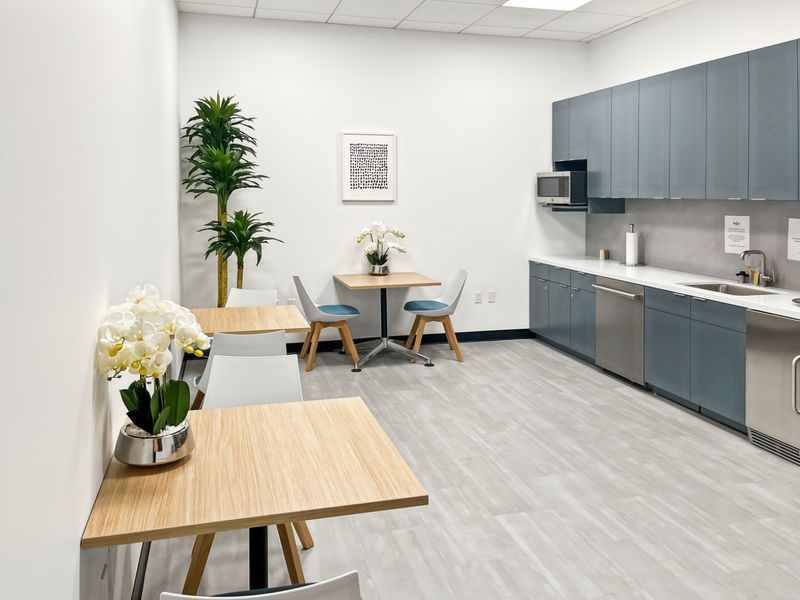 A modern office kitchenette featuring sleek blue cabinetry, integrated appliances, a stainless steel sink, and durable light-toned flooring.
