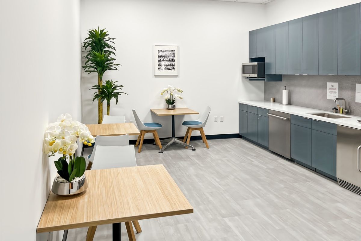 A modern office kitchenette featuring sleek blue cabinetry, integrated appliances, a stainless steel sink, and durable light-toned flooring.